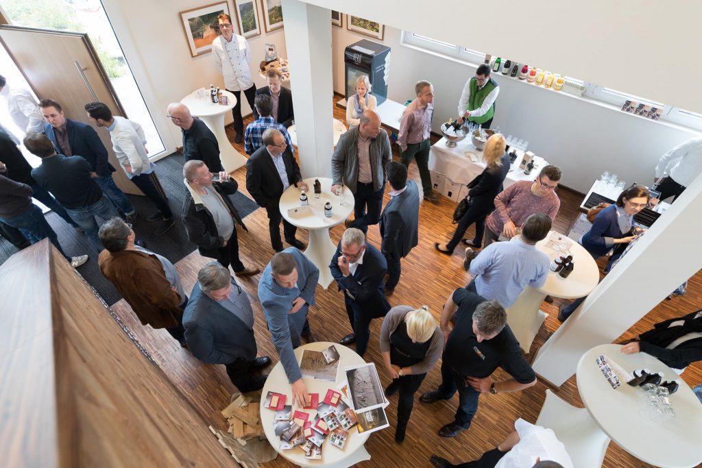 Foyer Coffee Consulate. Viele Personen stehen um Stehtische verteilt und unterhalten sich. 